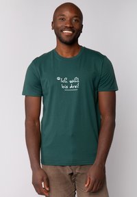 Homme souriant portant un T-shirt vert foncé avec du texte blanc en allemand, se tenant devant un fond clair.