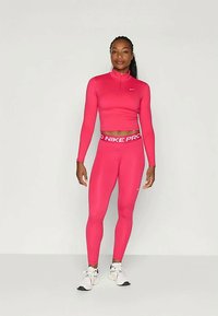 Tett rosa Nike Pro langermet topp med halvlukket krage, kombinert med matchende rosa leggings med en hvit logo belte. Hvite treningssko.