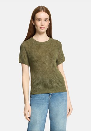 Femme aux longs cheveux roux portant un haut en maille à manches courtes couleur vert olive et un jean en denim bleu clair sur un fond uni.