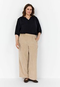 Blusa nera in tessuto leggero con scollatura a V e maniche corte, abbinata a pantaloni in corduroy beige a gamba larga e ballerine nere con dettagli dorati.