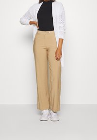 Cardigan en dentelle crème sur un top côtelé noir, associé à un pantalon large beige et des baskets blanches. Design simple, tissu texturé et couleurs neutres.