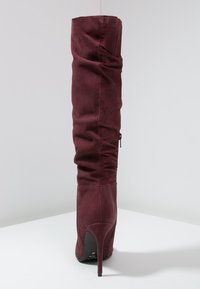 Bota de ante burdeos por encima de la rodilla con diseño arrugado, tacón de aguja alto y cremallera lateral; textura suave y forma ajustada.