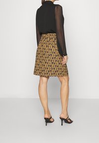 Blouse noire transparente à manches longues associée à une jupe marron longueur genou avec un motif de feuilles noires, complétée par des sandales à talons hauts noires.