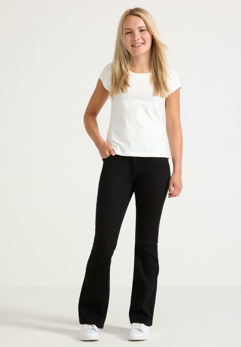 Vit kortärmad skjorta i kombination med svarta utsvängda jeans och vita sneakers. Modellen står med en hand i fickan och ler mot kameran.