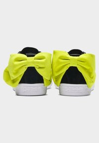Baskets Puma noires avec de grands nœuds en tissu jaune vif attachés aux talons, vues de dos sur un fond blanc.