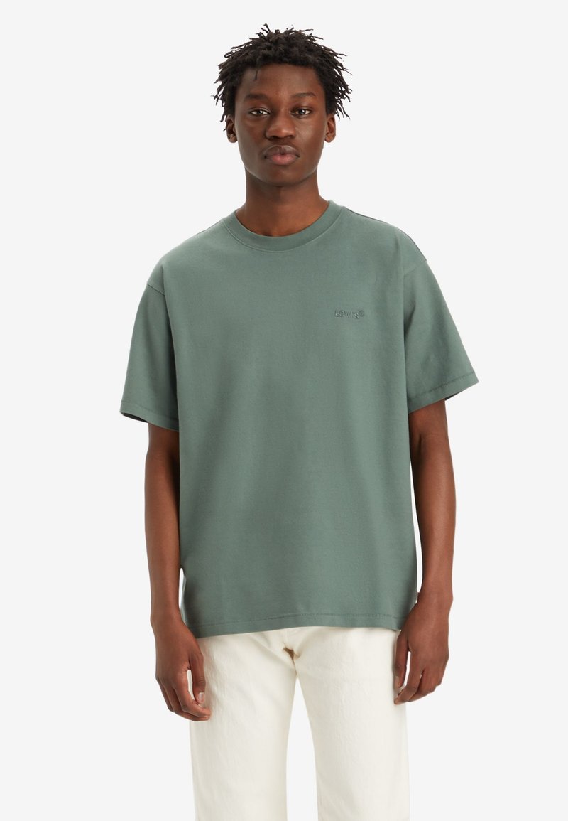Grünes Baumwoll-T-Shirt mit kurzen Ärmeln, Rundhalsausschnitt und minimaler Nahtverarbeitung. Verfügt über ein kleines, erhabenes Logo auf der Brust. Kombiniert mit weißen Hosen.