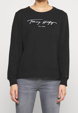 Woman wearing black long-sleeve Tommy Hilfiger sweatshirt with white logo and gray jeans, standing against a plain background.