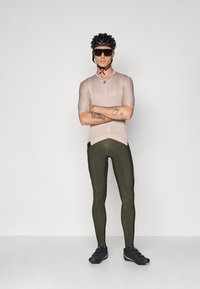 Tenue de cyclisme comprenant un maillot à manches courtes rose clair avec une fermeture éclair et un collant vert foncé. Les accessoires incluent un casque noir et des lunettes de soleil.