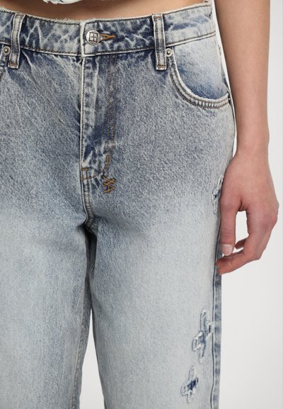 Blue denim jeans feature a relaxed fit, light wash with fading, subtle distressing, and embroidered floral accents on the thigh.