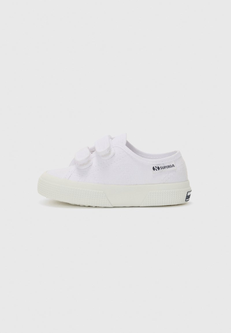 Zapatillas de lona blancas con punta redonda, dos correas de velcro y suela de goma texturizada; presenta una marca en el costado.