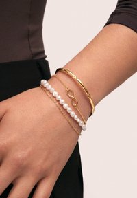 Brazalete de oro, pulsera de perlas con cadena de oro y un charm de infinito en una muñeca. Presenta texturas suaves y un diseño simple y elegante.
