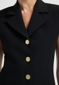 Zwarte mouwloze blazer met brede revers en drie grote ronde gouden knopen aan de voorkant, gedragen door een persoon.