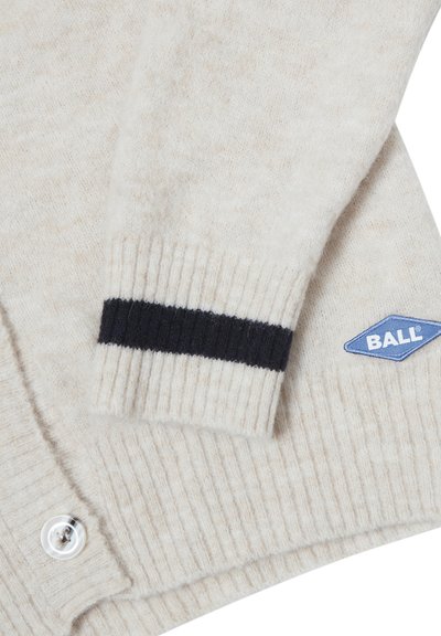 Manche de cardigan en maille crème avec une bande noire au niveau du poignet et un patch bleu en forme de diamant portant l'inscription « BALL », replié sur un corps côtelé avec un bouton transparent.