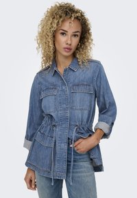 Veste utilitaire en denim avec un cordon de serrage à la taille, deux poches poitrine et des manches retroussées. Couleur bleu clair avec une finition texturée.
