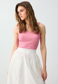Jeune femme aux cheveux bruns ondulés portant un haut rose sans manches et une jupe blanche taille haute, regardant sur le côté sur un fond clair.