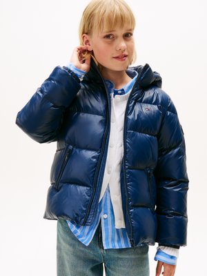 Navyblaue Steppjacke mit glänzender Oberfläche, hohem Kragen, Reißverschlusstaschen und Kapuze, kombiniert mit einem weißen Hemd und einem blauen gestreiften Oberteil.