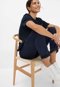 Marineblauwe t-shirt met korte mouwen, marineblauwe broek en witte sneakers. Zittend op een houten stoel met een geweven zitting. Natuurlijk licht.