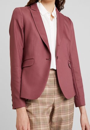 Kvinde iført rosa tilpasset blazer over hvid skjorte med krave og beige ternede bukser mod grå baggrund.
