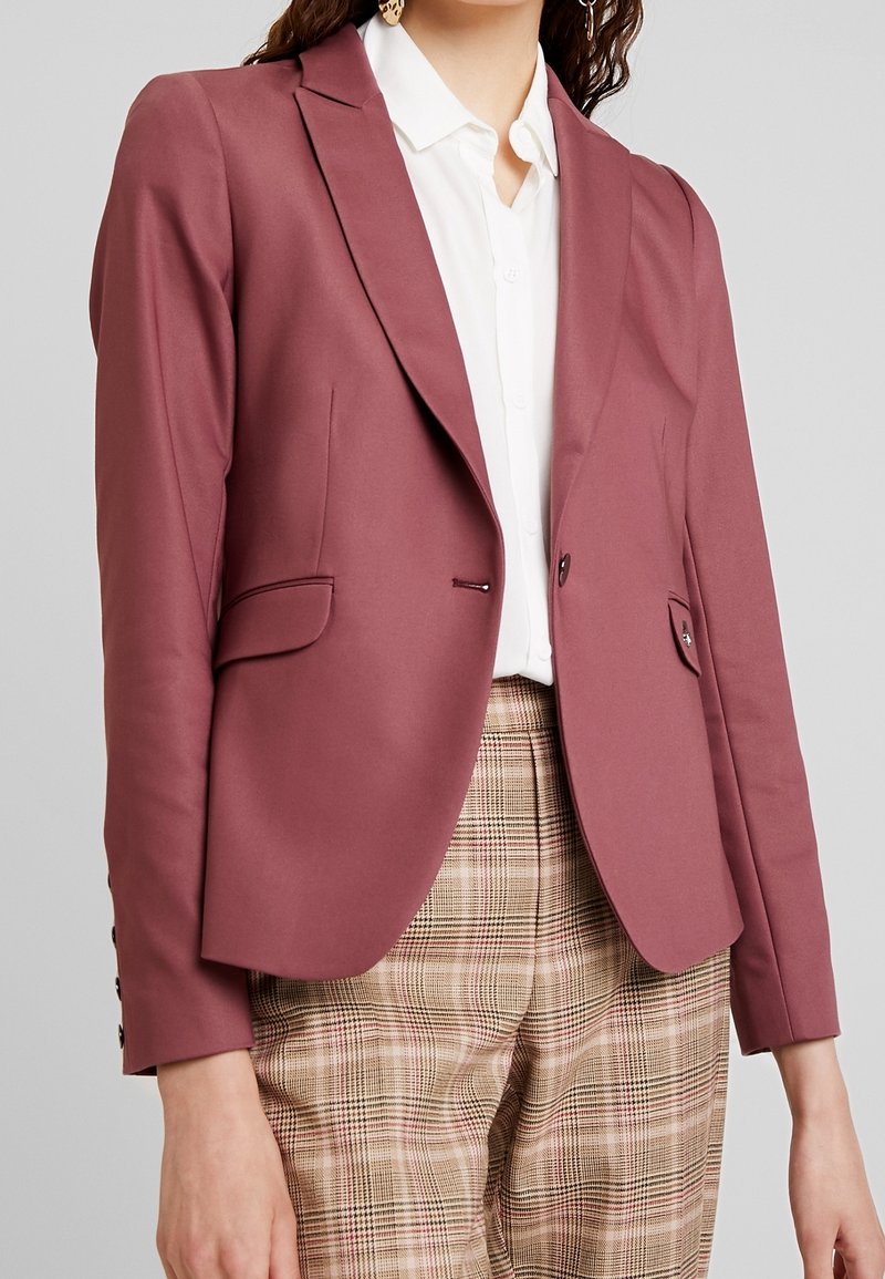 Femme portant un blazer rose sur mesure par-dessus une chemise blanche à col avec un pantalon beige à carreaux sur fond gris.