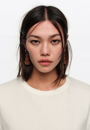 Young woman with straight dark hair wearing brown teddy bear earrings and a white crew neck top, facing forward with neutral expression.