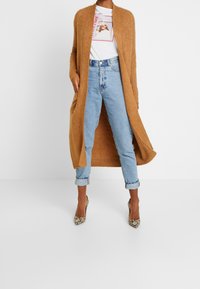 En person bär en lång tanfärgad cardigan över en vit t-shirt med grafik och blå jeans, i kombination med klackar med ormskinnsmönster, står mot en vit bakgrund.