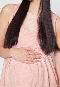 Ljusrosa ärmlös graviditetstopp med lös passform, samlad design vid midjan och slät textur, bärs med händerna vilande på magen.