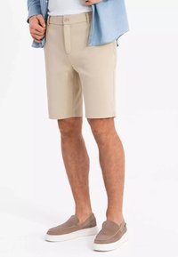 Pantaloni corti beige con una texture liscia, dotati di chiusura frontale con bottone, tasche laterali e una vestibilità sartoriale, abbinati a scarpe slip-on marroni.