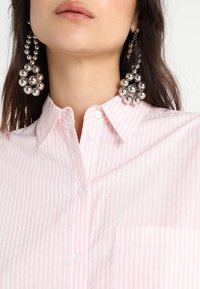 Camicia button-up a righe rosa chiaro con taschino frontale, abbinata a orecchini pendenti con grandi perline d'argento che presentano un design a grappolo.