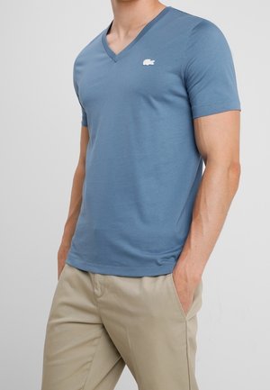 Uomo che indossa una maglietta blu a maniche corte con scollo a V e logo bianco sul petto, e pantaloni beige, con le mani nelle tasche.