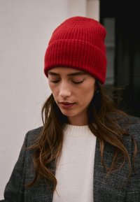 Red ribbed knit beanie with a fold-over cuff, worn with a white sweater and gray blazer. Soft texture with a close fit on the head.