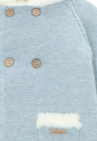 Abrigo tejido en azul claro con cuello forrado de piel, botones de madera y un bolsillo acolchado con una etiqueta de madera que dice "Aranda".