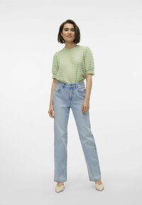 Ανοιχτό πράσινο υφαντό μπλούζα κοντομάνικη συνδυασμένη με ψηλόμεσο ανοιχτό μπλε τζιν κατ' ευθείαν και μπεζ γόβες. Απλό λευκό φόντο.