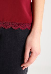 Bordeauxfärgad topp med spetsdetaljer i nederkant, som har en slät textur. Hand vilande mot mörk denim, vilket framhäver plagget detaljer.