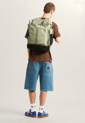Zielony plecak z tkaniny z wieloma kieszeniami, czarną dolną częścią i wyściełanymi paskami. Model nosi brązową koszulę i niebieskie dżinsowe szorty.