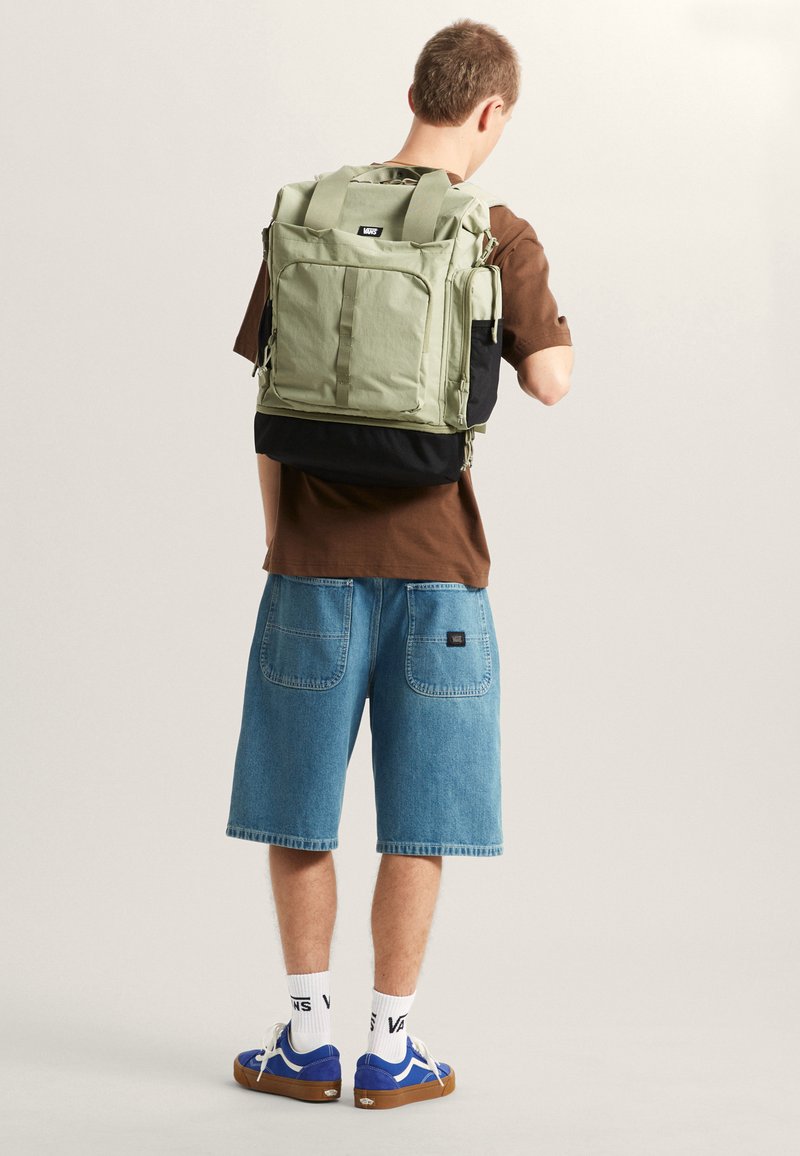 Sac à dos en tissu vert avec plusieurs poches, partie inférieure noire et bretelles rembourrées. Le modèle porte une chemise marron et un short en denim bleu.
