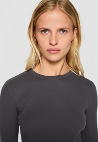 Tmavě šedý dlouhý rukáv top s vroubkovanou texturou a kulatým výstřihem. Přiléhavý design, hladký materiál, žádné viditelné vzory nebo akcenty.