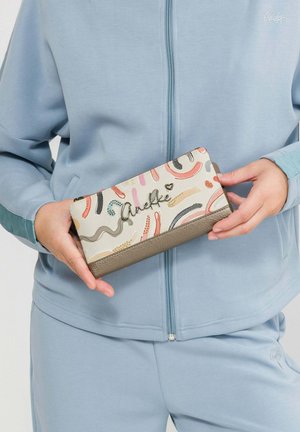 Hände halten eine mehrfarbige Geldbörse mit abstrakten Mustern und dem "Anekke"-Logo, Person trägt eine hellblaue Jacke mit Reißverschluss und passende Hose.