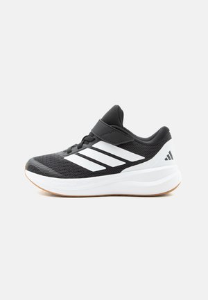Chaussure de sport noire et blanche avec trois bandes blanches, fermeture velcro, semelle blanche rembourrée et design en mesh respirant.
