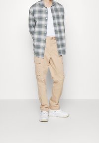 Grön och vit rutig långärmad skjorta över en vit linne, beige cargo-byxor med fickor och vita sneakers.
