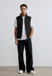 Νέος άνδρας στέκεται φορώντας μαύρο αμάνικο γιλέκο, λευκό μπλουζάκι, σκούρα τζιν με φαρδιές παντελόνες και λευκά αθλητικά παπούτσια, σε απλό φόντο.