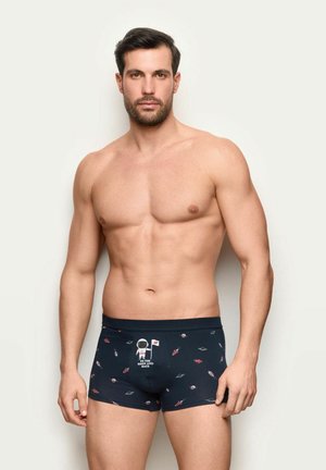 Muskularny mężczyzna z ciemnymi włosami, ubrany w granatowe bokserki zdobione astronautami, rakietami, planetami oraz napisem "Na księżyc i z powrotem".