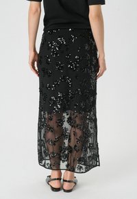 Jupe noire en sequins avec motifs floraux, superposition transparent, et design mi-long, complétée par une taille ajustée et des sandales noires élégantes.
