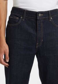 Jeans in denim blu scuro con taglio dritto, cinque tasche, cuciture visibili di colore giallo a contrasto e chiusura a bottoni.