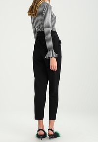 Svarta högmidjade byxor med en smal passform, kombinerade med en svartvit randig långärmad topp. Skorna har gröna fjäderdetaljer.