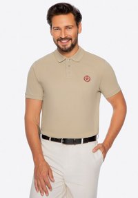 Beige polo tričko s texturovaným povrchem, vybavené límcem a třemi knoflíky, zdobené červeným vyšívaným logem. S kombinací bílých kalhot.