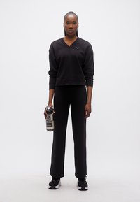 Γυναίκα που στέκεται μπροστά, φοράει μαύρα αθλητικά ρούχα Puma και αθλητικά παπούτσια, κρατώντας ένα διαφανές μπουκάλι νερού Puma στο δεξί της χέρι.