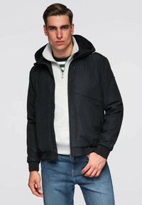 Czarna kurtka z zamkiem i kapturem, zaprojektowana z gładkiej tkaniny, z dwiema przednimi kieszeniami i ściągaczami na mankietach. Noszona na szarym swetrze i dżinsach.