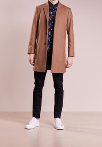Ruskea villakangas takki, jossa on muotoutunut design, kaksi nappia, yhdistetty tumman kukkakuosisen paidan ja mustien housujen kanssa, täydentää valkoiset sneakerit.