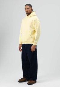 Geel pullover hoodie met een buidelzak, geribbelde boorden en subtiele branding, gecombineerd met donkerblauwe jeans en bruine veterschoenen.