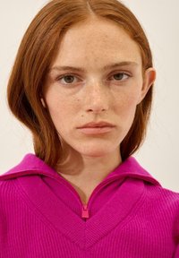 Fuksjowy sweter zapinany na zamek o strukturalnej teksturze, z szerokim dekoltem w kształcie litery V i różowym detalem zamka przy kołnierzu.